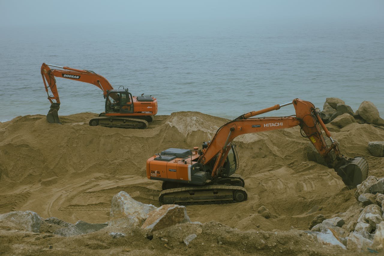 About Two excavators operate at a sandy construction site by the sea, moving sand and rock.